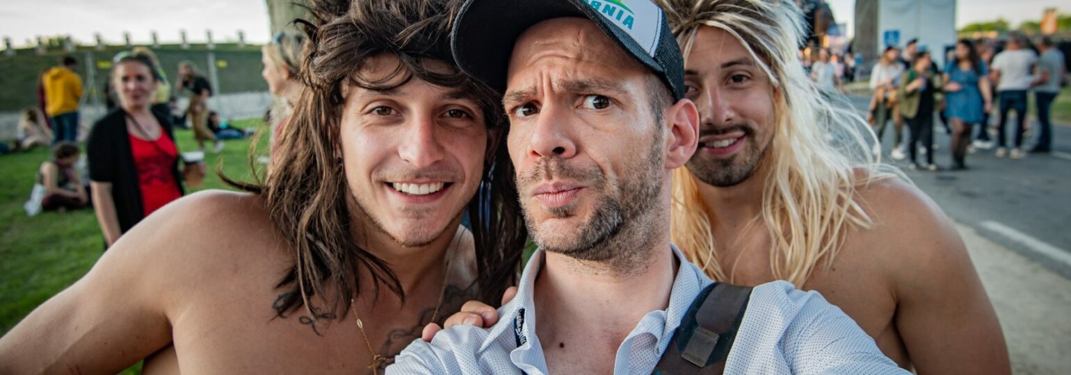 Trois hommes souriants prennent un selfie en extérieur.