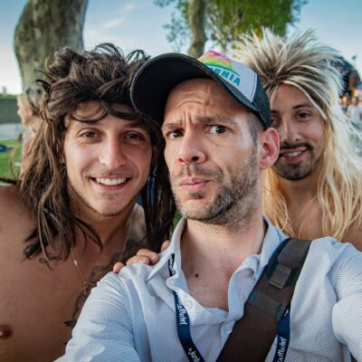 Trois hommes souriants prennent un selfie en extérieur.