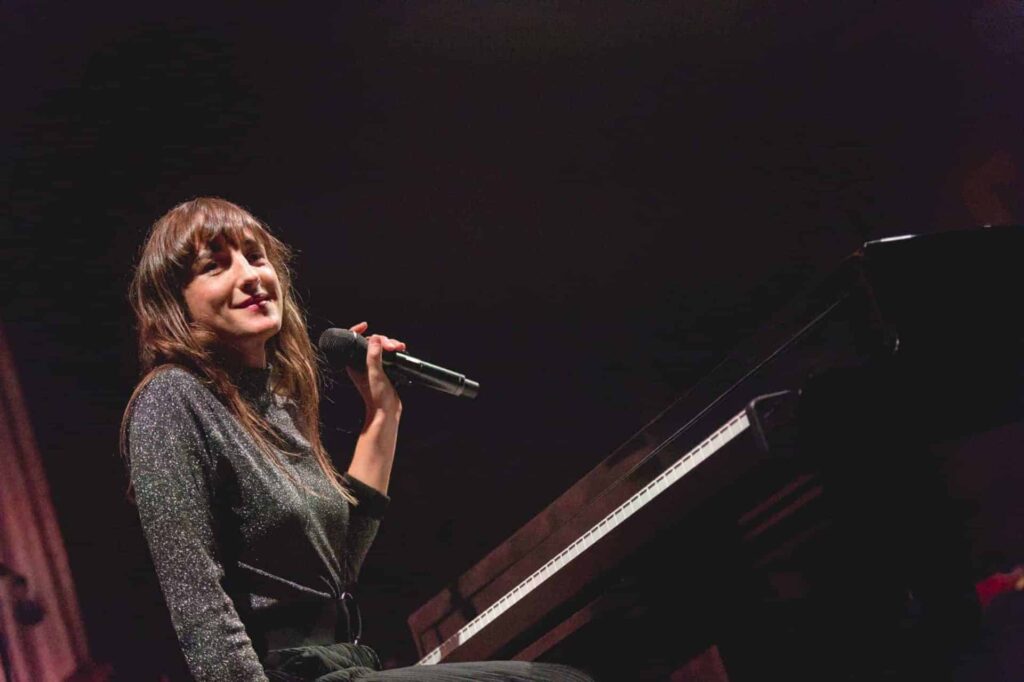 Femme souriant tenant un microphone, assise au piano.