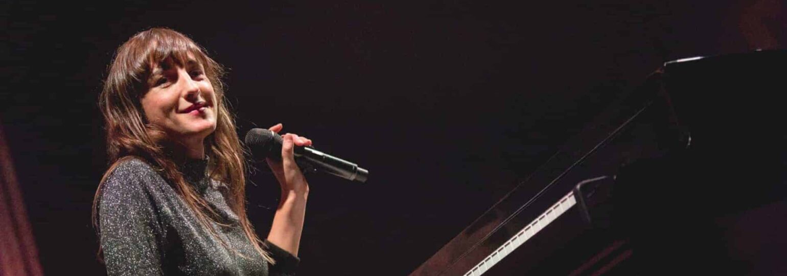 Femme souriant tenant un microphone, assise au piano.