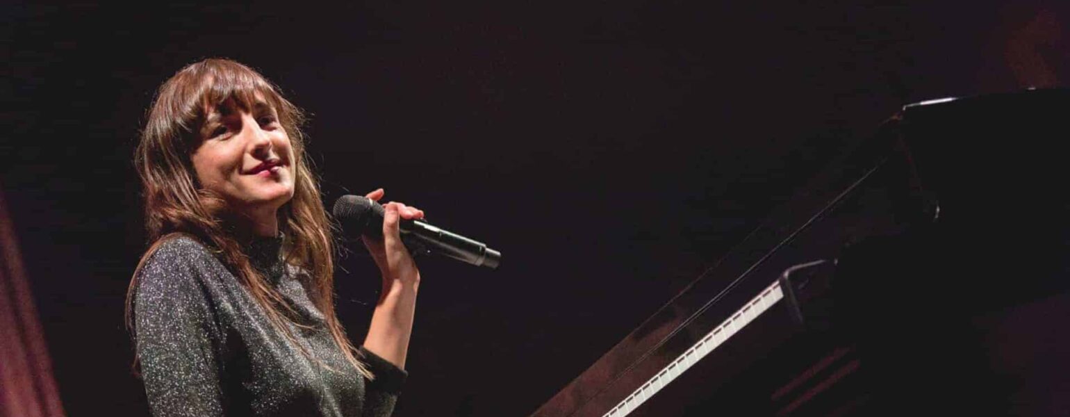 Femme souriant tenant un microphone, assise au piano.