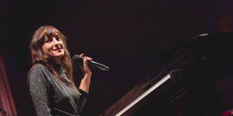 Femme souriant tenant un microphone, assise au piano.