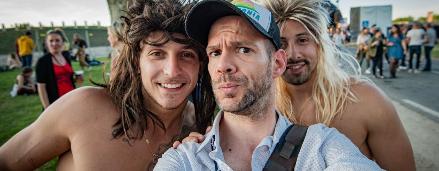 Trois hommes souriants prennent un selfie en extérieur.