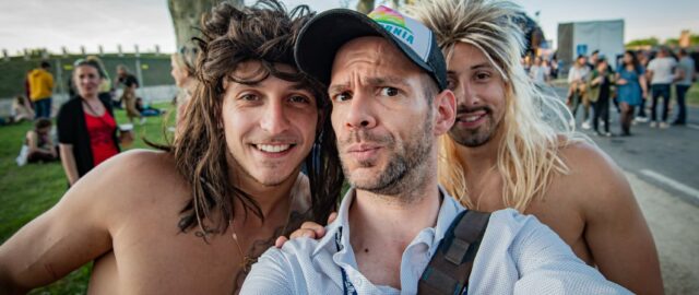 Trois hommes souriants prennent un selfie en extérieur.