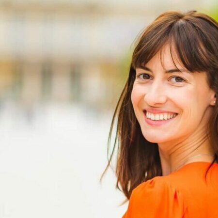 Femme souriante en extérieur flou, vêtue d'orange.
