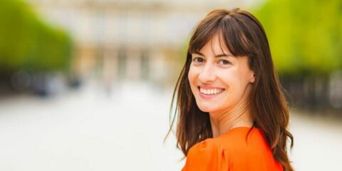 Femme souriante en extérieur flou, vêtue d'orange.
