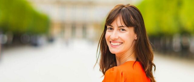 Femme souriante en extérieur flou, vêtue d'orange.