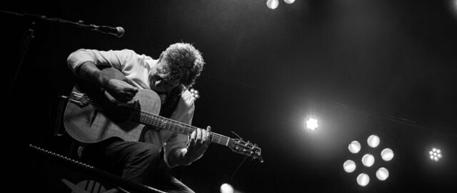 Musicien jouant de la guitare sur scène en noir et blanc.