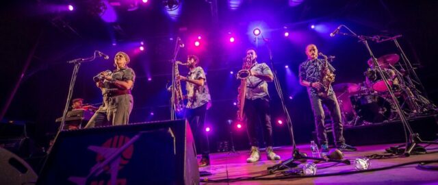 Groupe de musiciens en concert avec éclairage violet.