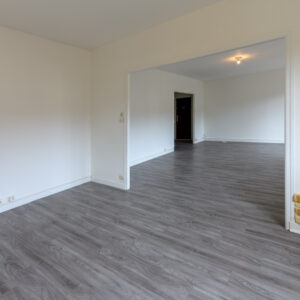 Salon vide avec parquet gris et mur en pierre.