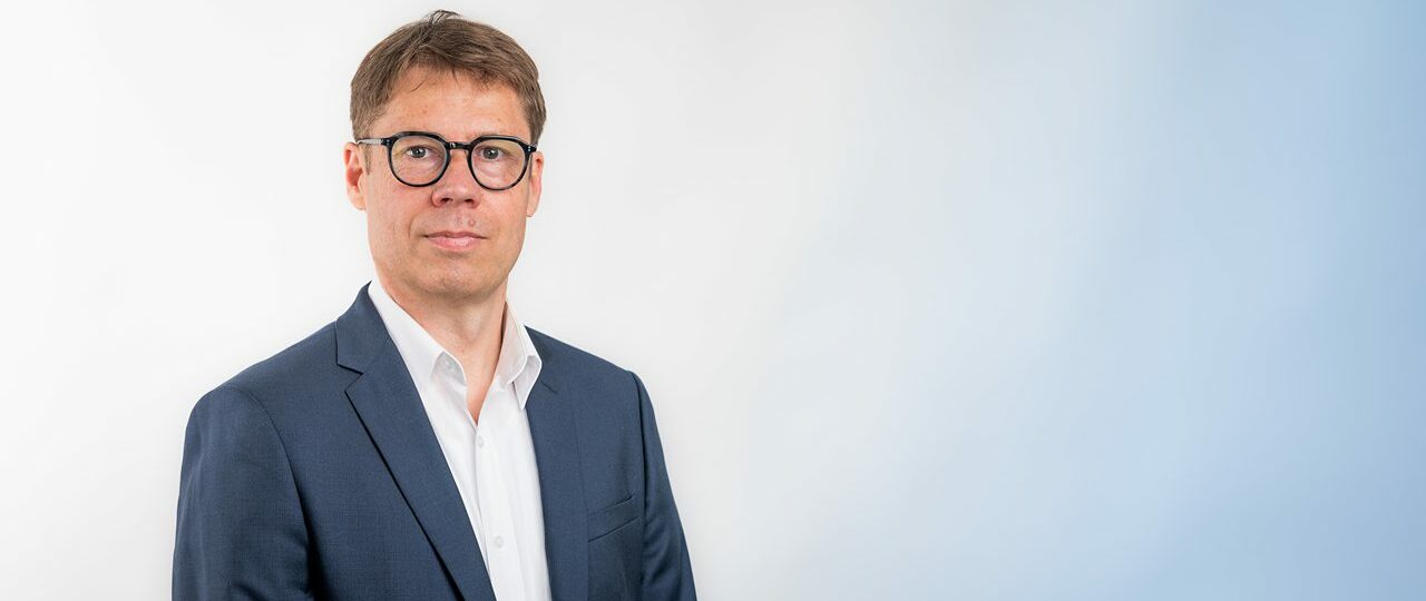 Homme en costume, lunettes noires, fond blanc-bleu.