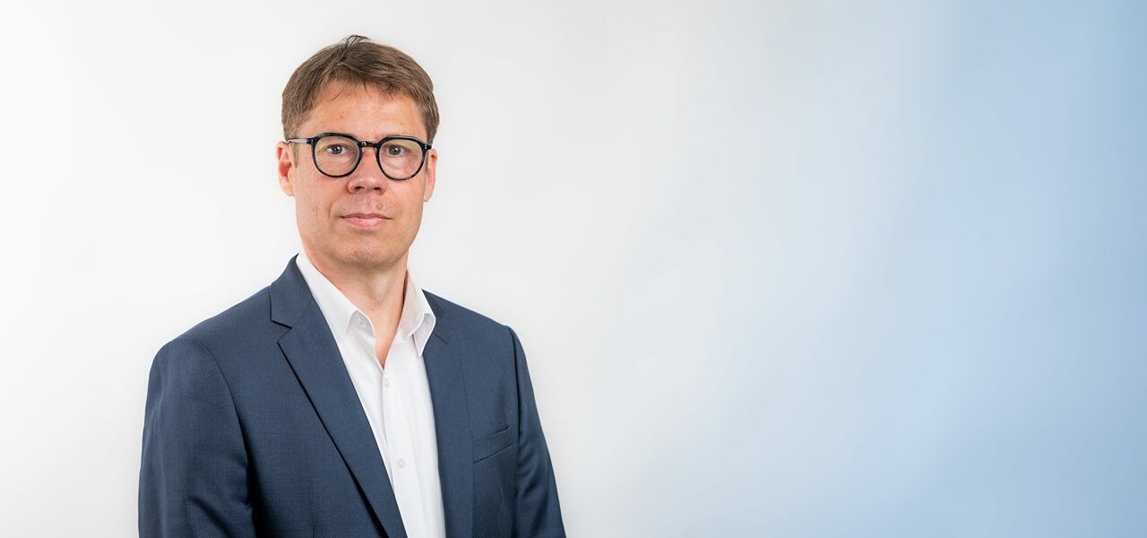 Homme en costume, lunettes noires, fond blanc-bleu.