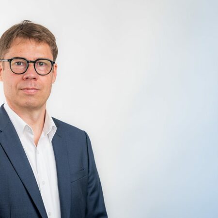 Homme en costume, lunettes noires, fond blanc-bleu.
