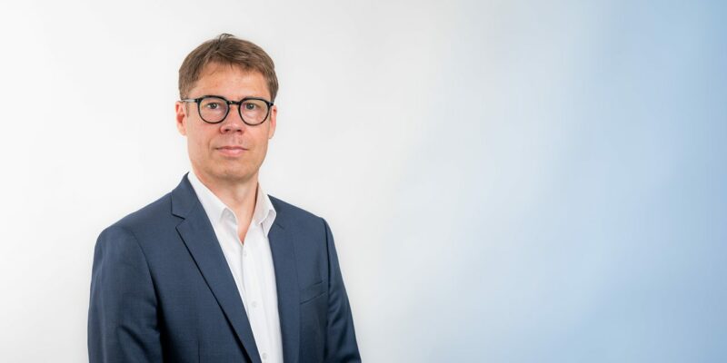 Homme en costume, lunettes noires, fond blanc-bleu.