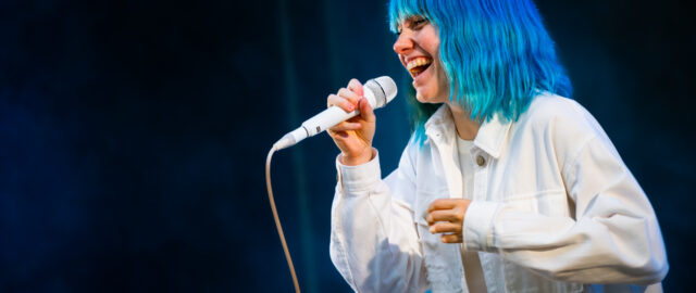 Personne chantant sur scène, cheveux bleus fluorescent.