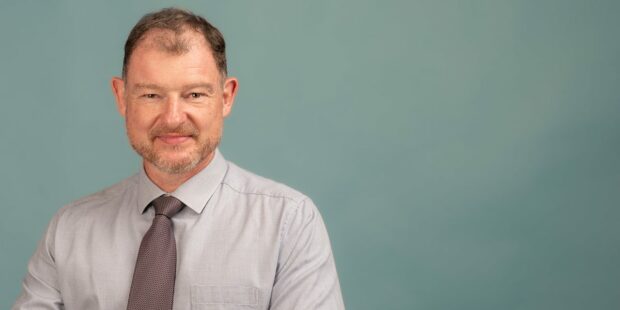 Homme souriant en chemise et cravate devant fond bleu.