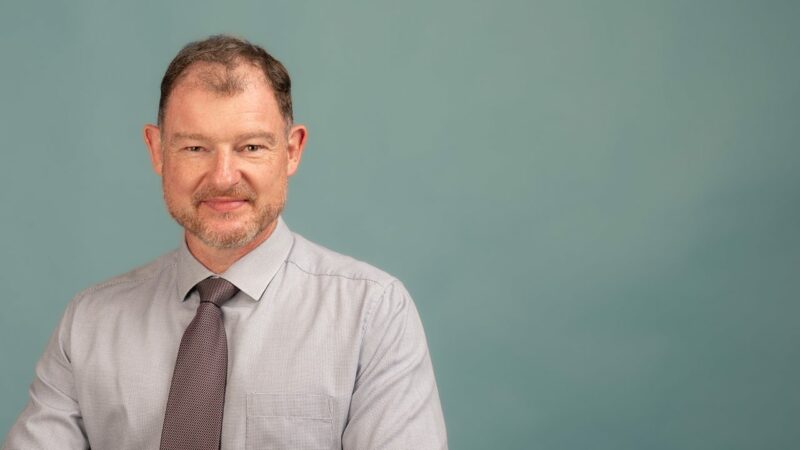 Homme souriant en chemise et cravate devant fond bleu.