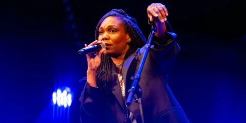 Chanteuse avec dreadlocks tenant un micro sur scène éclairée.