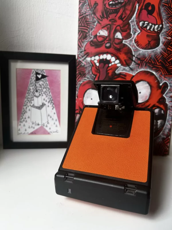 Appareil photo instantané orange devant des illustrations artistiques.