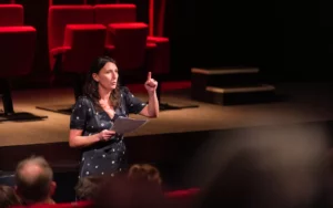 Femme s'exprimant sur scène devant des fauteuils rouges.